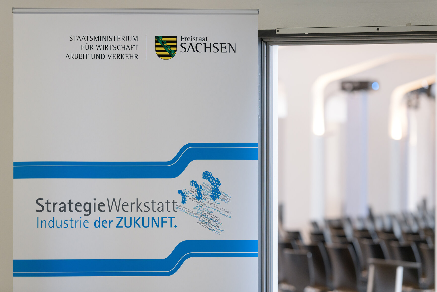 Ein Roll-Up mit dem Text »StrategieWerkstatt Industrie der Zukunft« hängt neben einer offenen Tür.