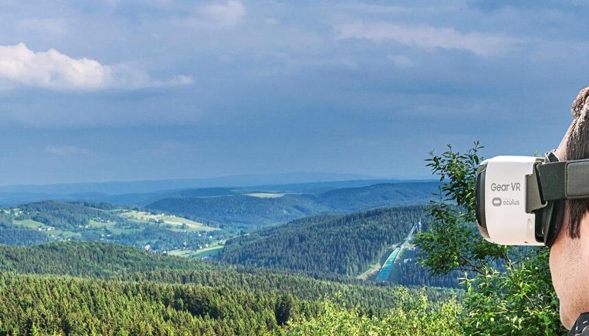 Fotomontage: ein Blick auf eine grüne bewaldete Landschaft, im rechten Bildrand ist der Kopf eines Menschen zu sehen, der mit einer Computerbrille in diese Landschaft schaut.