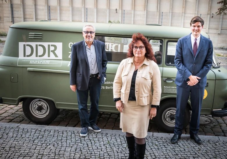 Zwei Männer und eine Frau stehen vor einen Matt-grünen Transporter