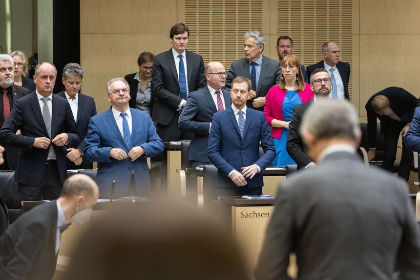 EIne Gruppe Menschen steht in einem Plenarsaal zu einer Trauerminute.